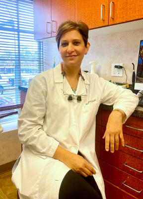 The image shows a woman seated in a dental office, wearing a white lab coat and smiling at the camera.