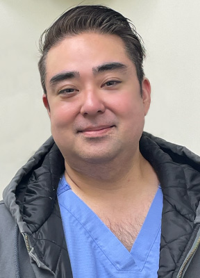 The image is a photograph of a man in a medical uniform, smiling and looking directly at the camera.