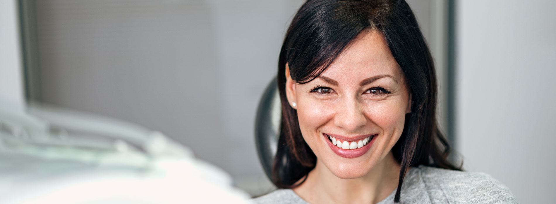 The image shows a smiling woman with dark hair, wearing a light-colored top, seated in an office setting with a glass partition and a blurred background.