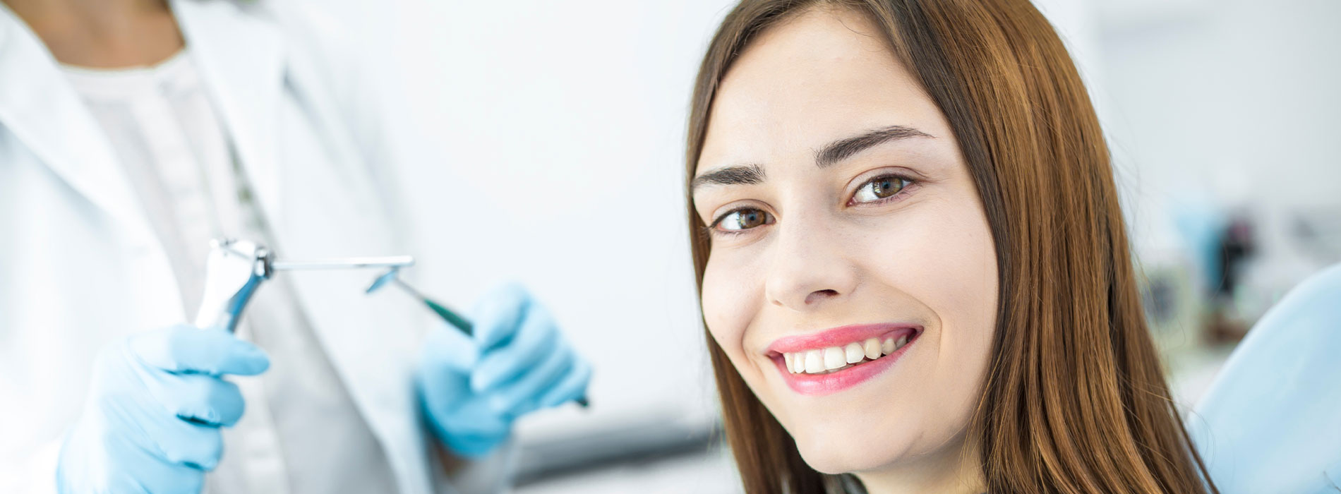 The image is a photograph featuring a smiling woman in the foreground, wearing makeup and a blue top, with a dental professional or hygienist in the background, dressed in a white lab coat and holding dental instruments.