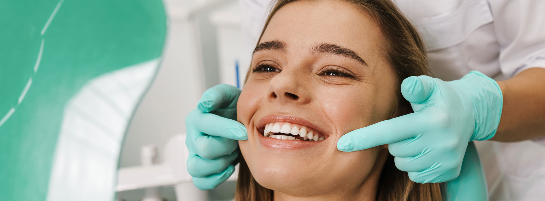 A woman seated in a dental chair, smiling as a dentist adjusts her mouth with their hands.