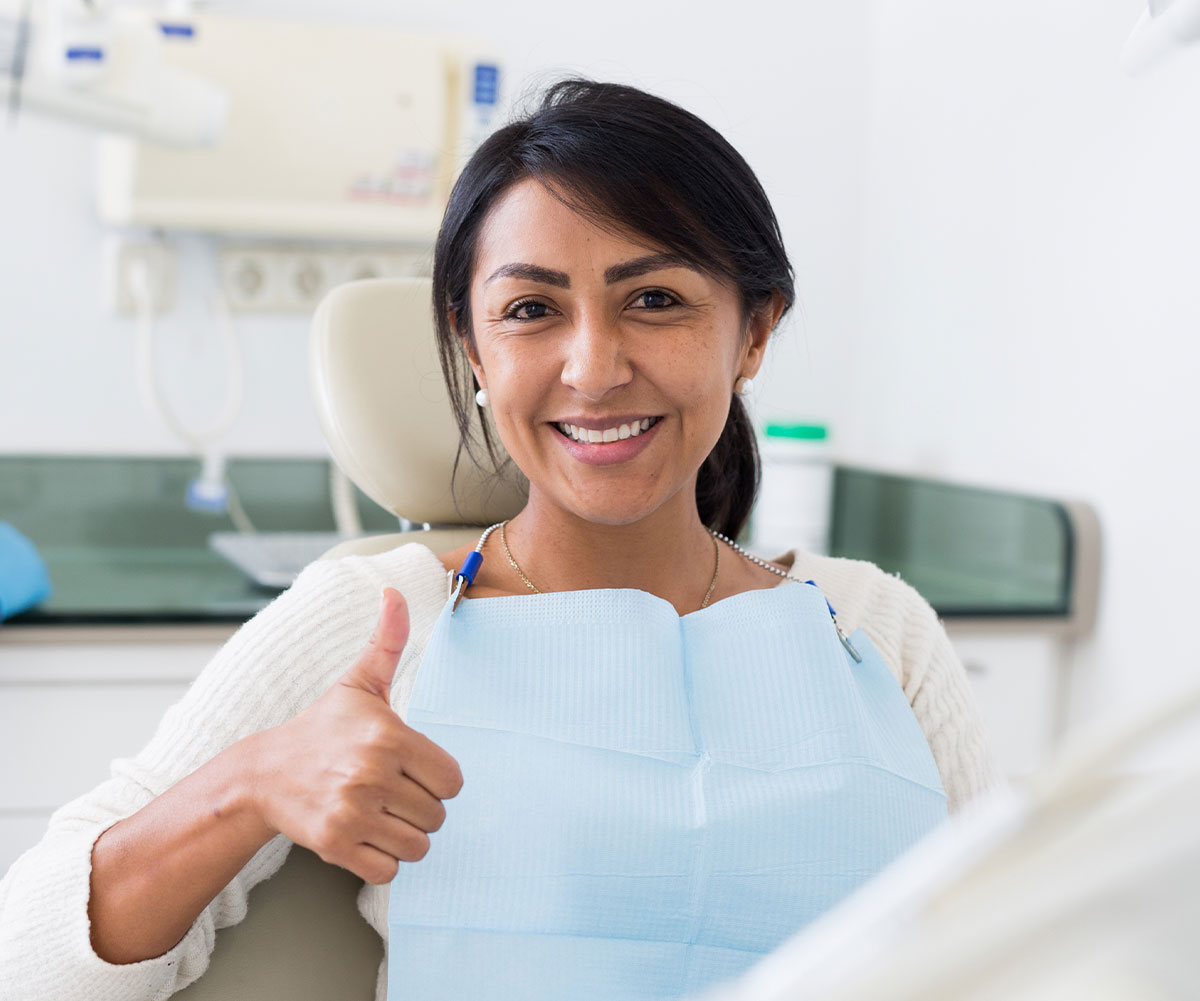 The image shows a person giving a thumbs-up gesture while sitting in a dental chair, smiling at the camera.