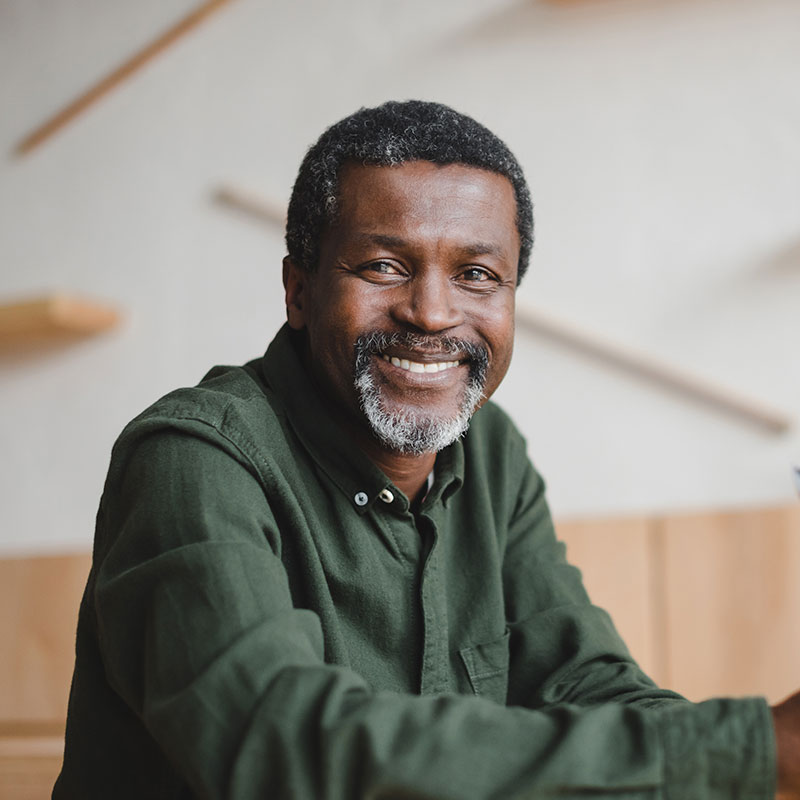 The image shows a man with gray hair, wearing a green shirt and a beard, sitting at a table with a wooden surface. He is smiling and appears to be in a positive mood.