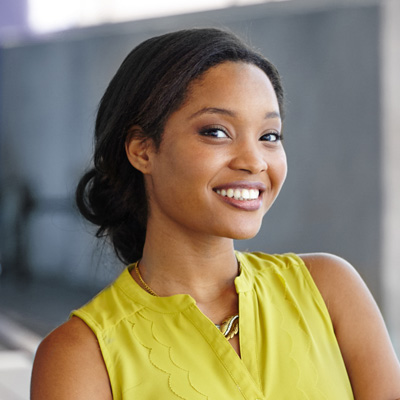 The image shows a smiling woman with dark hair, wearing a yellow top and standing against a blurred background.