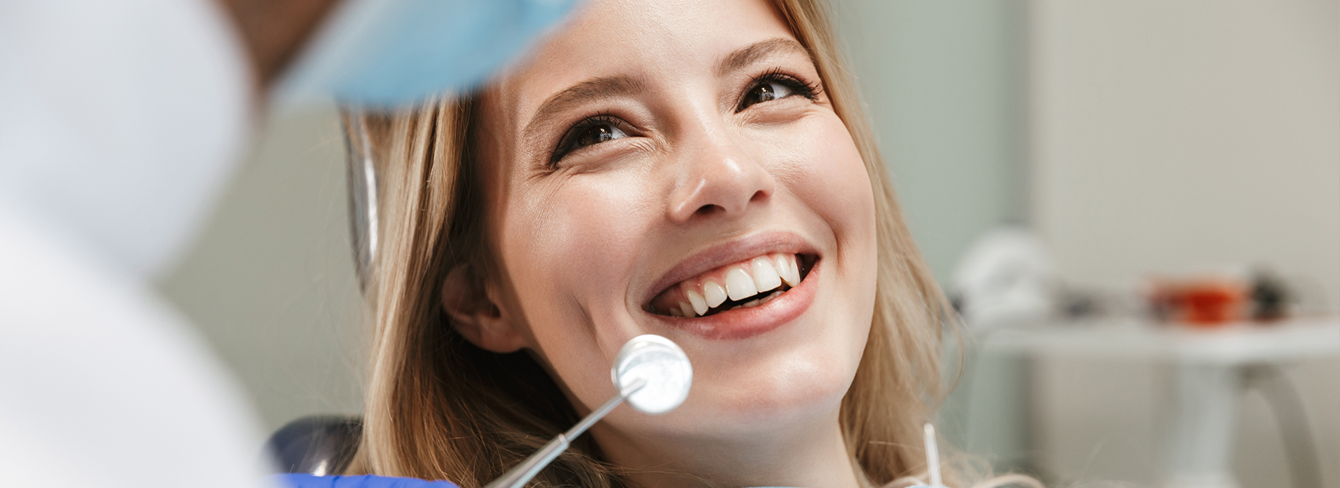 The image shows a woman smiling at the camera while being in a dental or medical setting, with a person wearing a white coat and cap visible in the background.