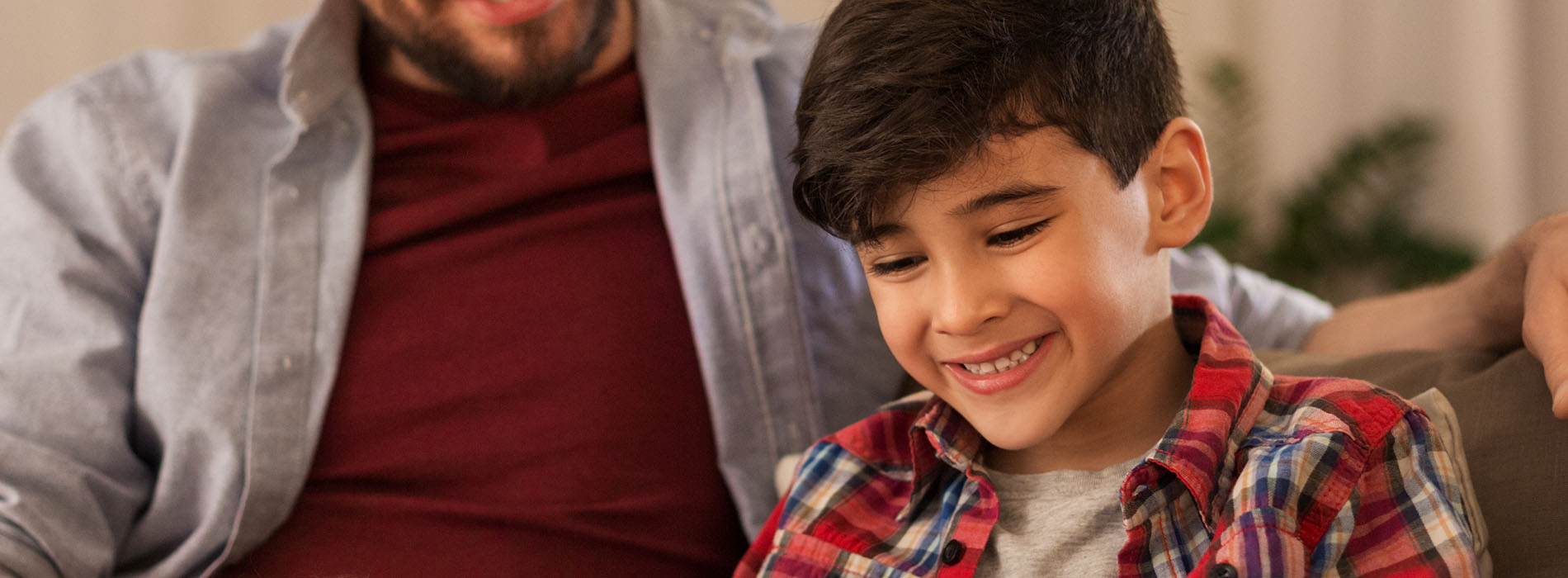 The image depicts a man and a child sitting on a couch, looking at something off-camera with smiles on their faces.