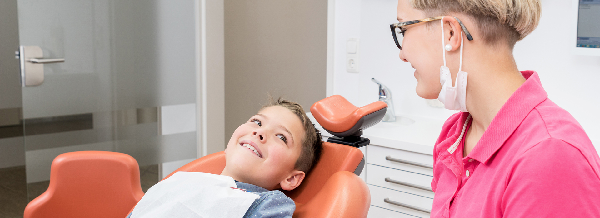 An image of a dental office with a dentist and patient, featuring a young child being checked by the dentist.