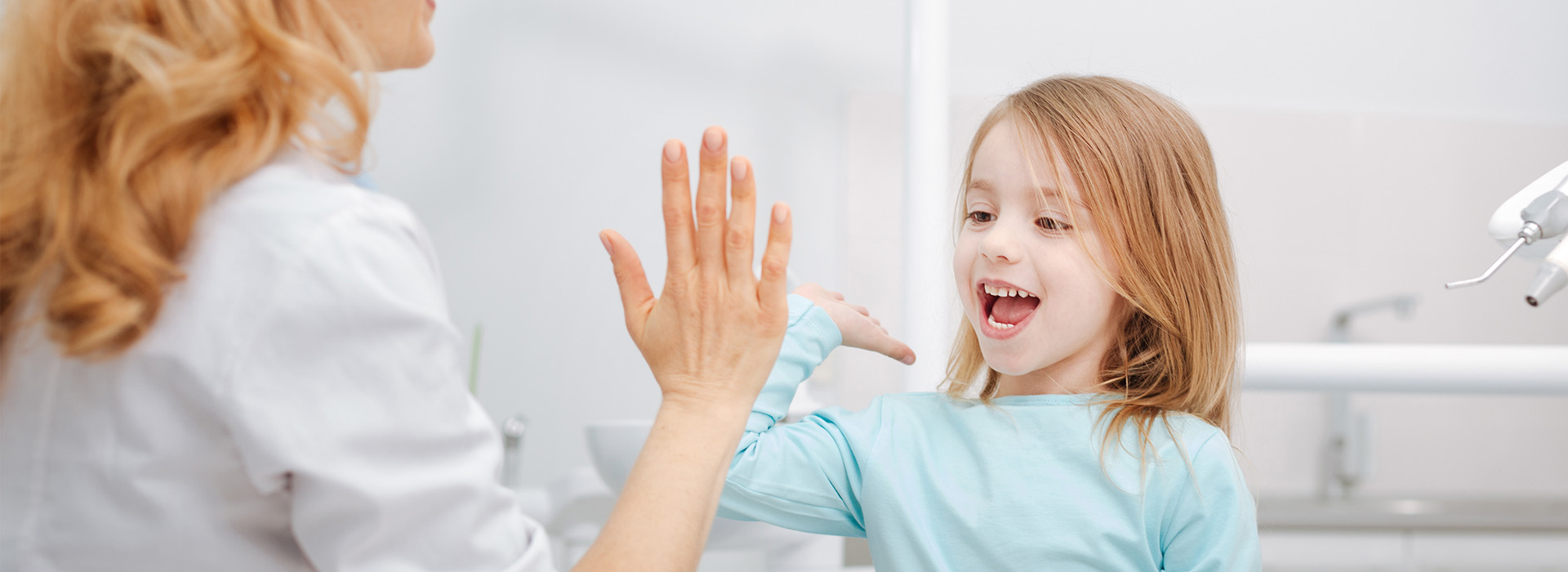 A woman and a young girl are seen in the image, with the woman holding up her hand towards the child. They appear to be indoors, possibly in a dental or medical setting given the presence of a sink and what seems like a dental chair in the background. The woman is smiling at the camera while the child looks away.