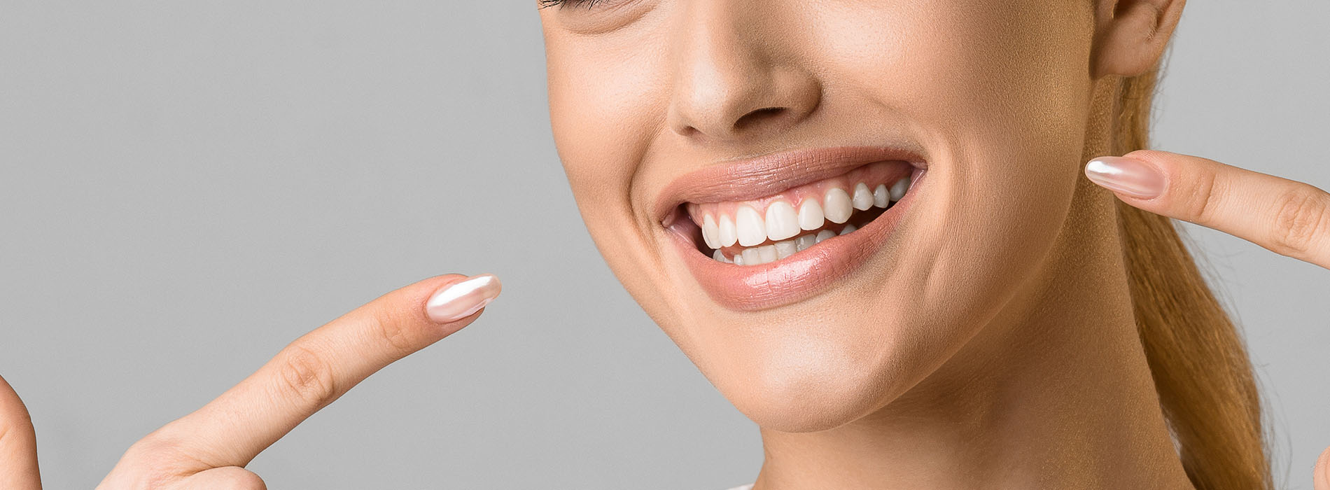 A smiling woman with a manicured hand gesturing towards her teeth, set against a neutral background.