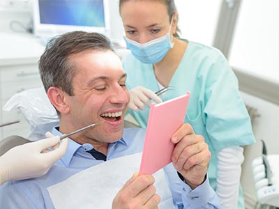 The image shows a man in a dental chair, holding up a pink card with a smile on his face, while a dentist and hygienist are working behind him.