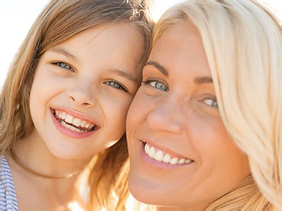 A woman and a young girl are smiling at the camera, with the woman appearing to be the mother of the child. They are standing close together outdoors during daylight, likely on a sunny day given the bright lighting.