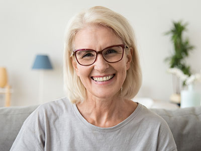 The image features a woman with short blonde hair, wearing glasses and a light-colored shirt, smiling at the camera. She appears to be indoors, possibly in a living room setting, as suggested by the presence of a sofa and a lamp in the background.