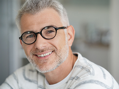 The image is a photograph of a man with gray hair and glasses, smiling slightly towards the camera. He has a beard and mustache, and he appears to be indoors, possibly in an office or home setting.