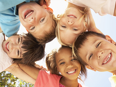 The image shows a group of children, likely at an outdoor event or gathering, with their arms around each other, smiling and posing for the camera.