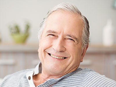 A smiling elderly man with white hair, wearing a dark shirt and blue jeans, sitting in a relaxed posture.