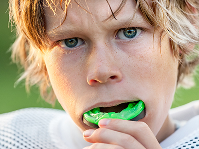 An image of a young boy with blonde hair, wearing a sports jersey and holding a green toothbrush in his mouth.