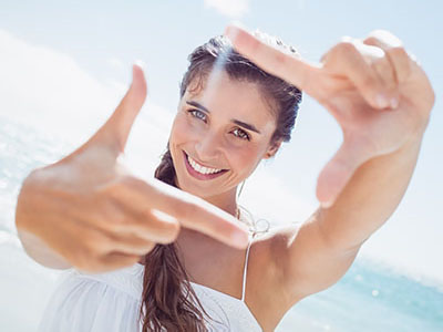 The image shows a woman with long hair, smiling at the camera while holding her hand up to create a frame around it. She is outdoors under bright sunlight, wearing a white top and sunglasses, with a clear sky in the background.